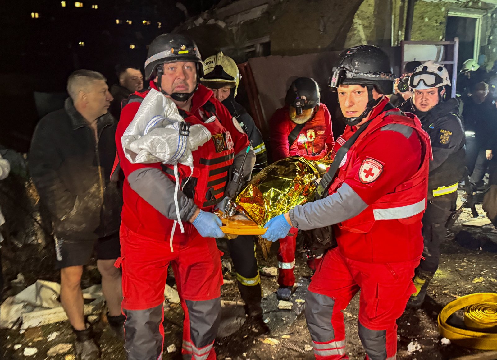 Emergency responders in red uniforms transporting a body wrapped in a thermal blanket on a stretcher at night