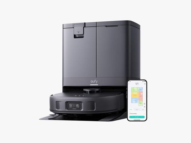 Black circular robotic vacuum docked in a tall black station. Small mobile phone to the right, displaying a floorplan.