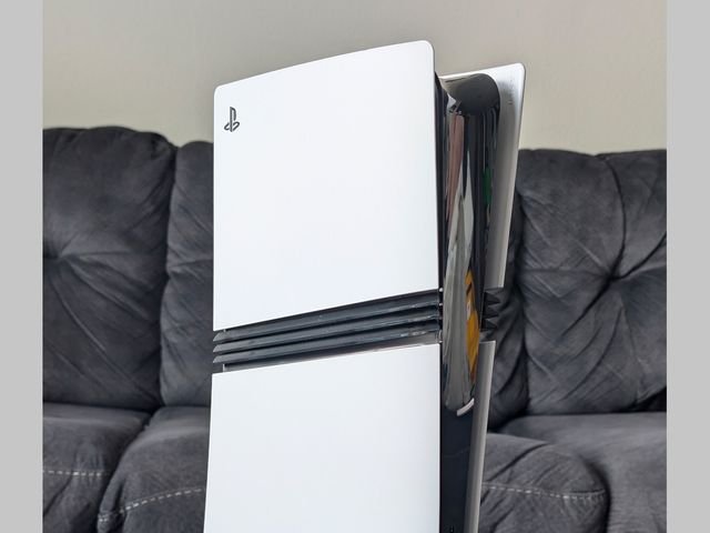 Front view of Sony PlayStation 5, a slim white video game console with flared top edge, sitting on a wooden surface