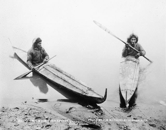 Historic black-and-white photo showing hunters paddling wooden kayaks along shore