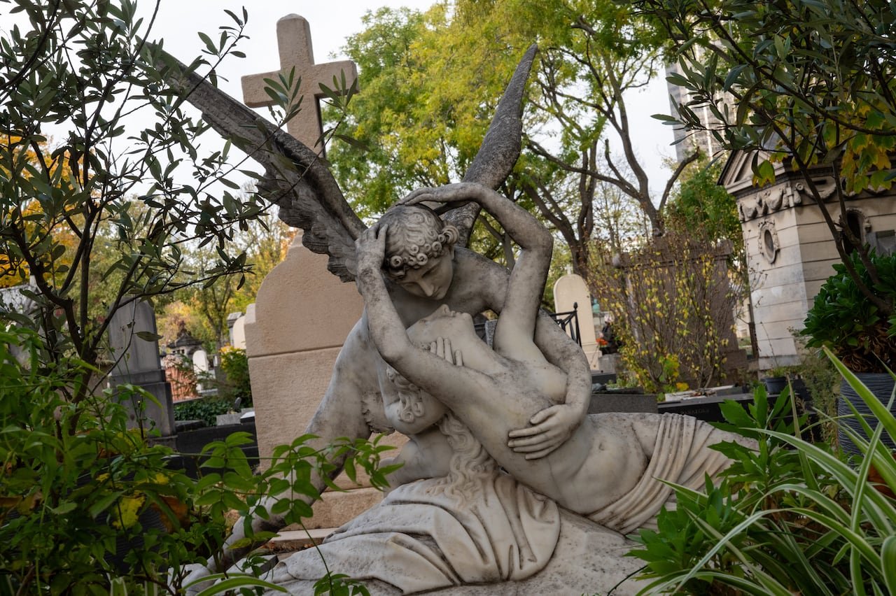 Angel statue embracing woman amid greenery at Montparnasse Cemetery