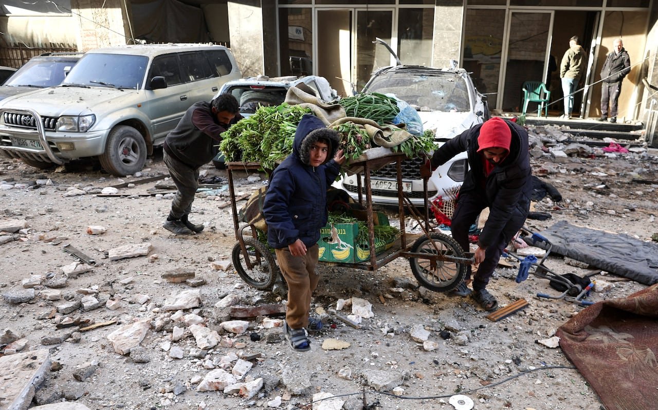 Two men and child pushing produce cart through rubble-strewn streets.