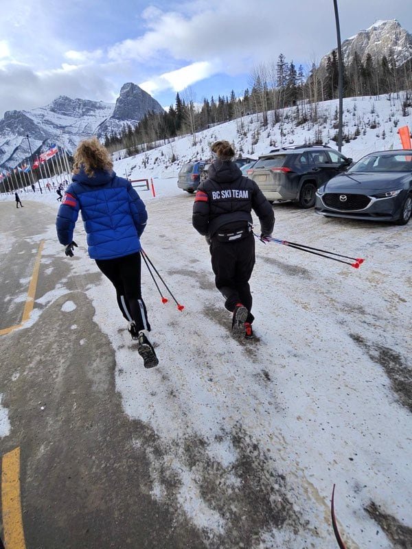 Two girls run together holding ski poles with mountains in background