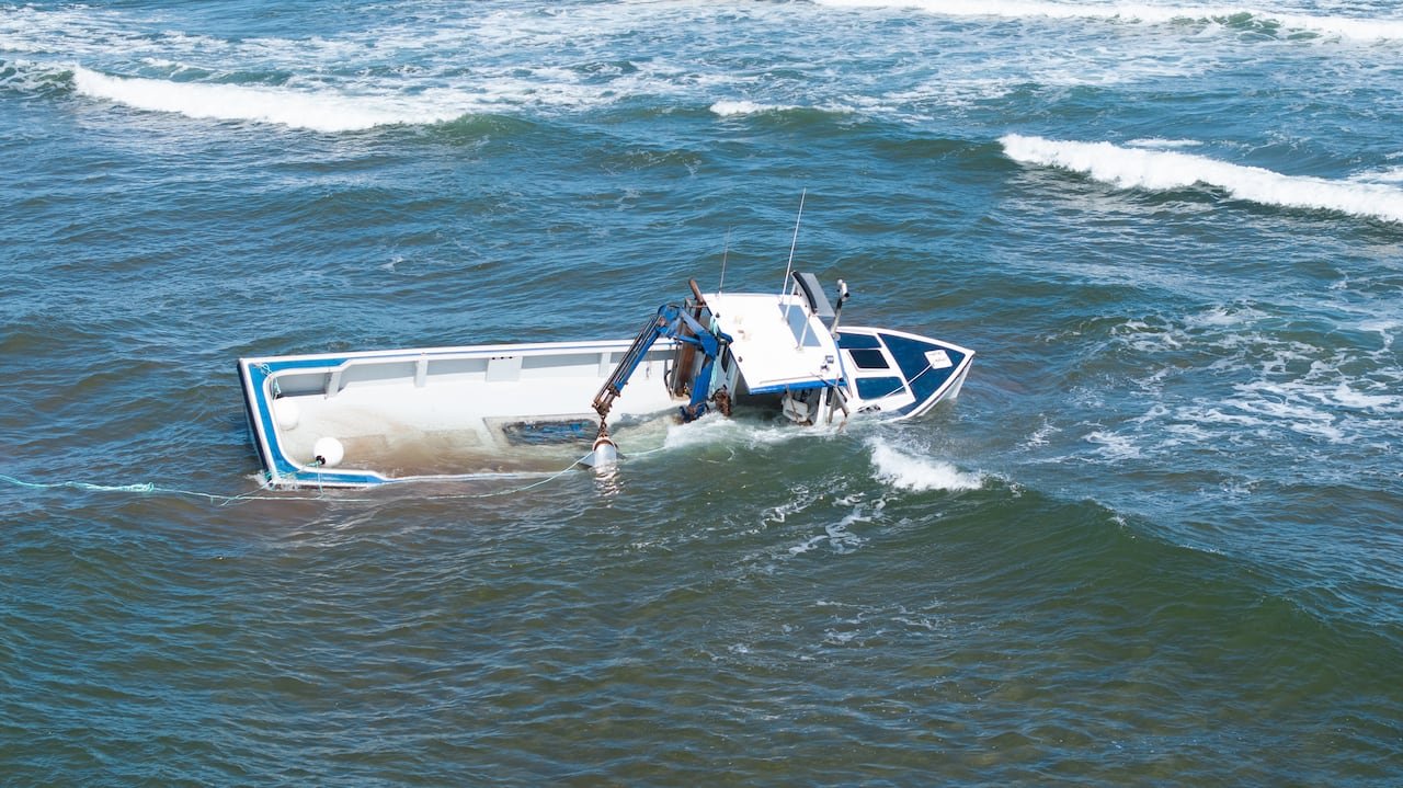 Aerial view showing partially submerged blue and white fishing boat near sandbar wiht waves crashing.