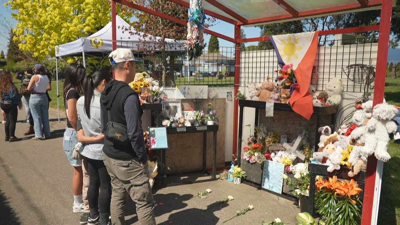 People standing solemnly before floral tributes under shelter.