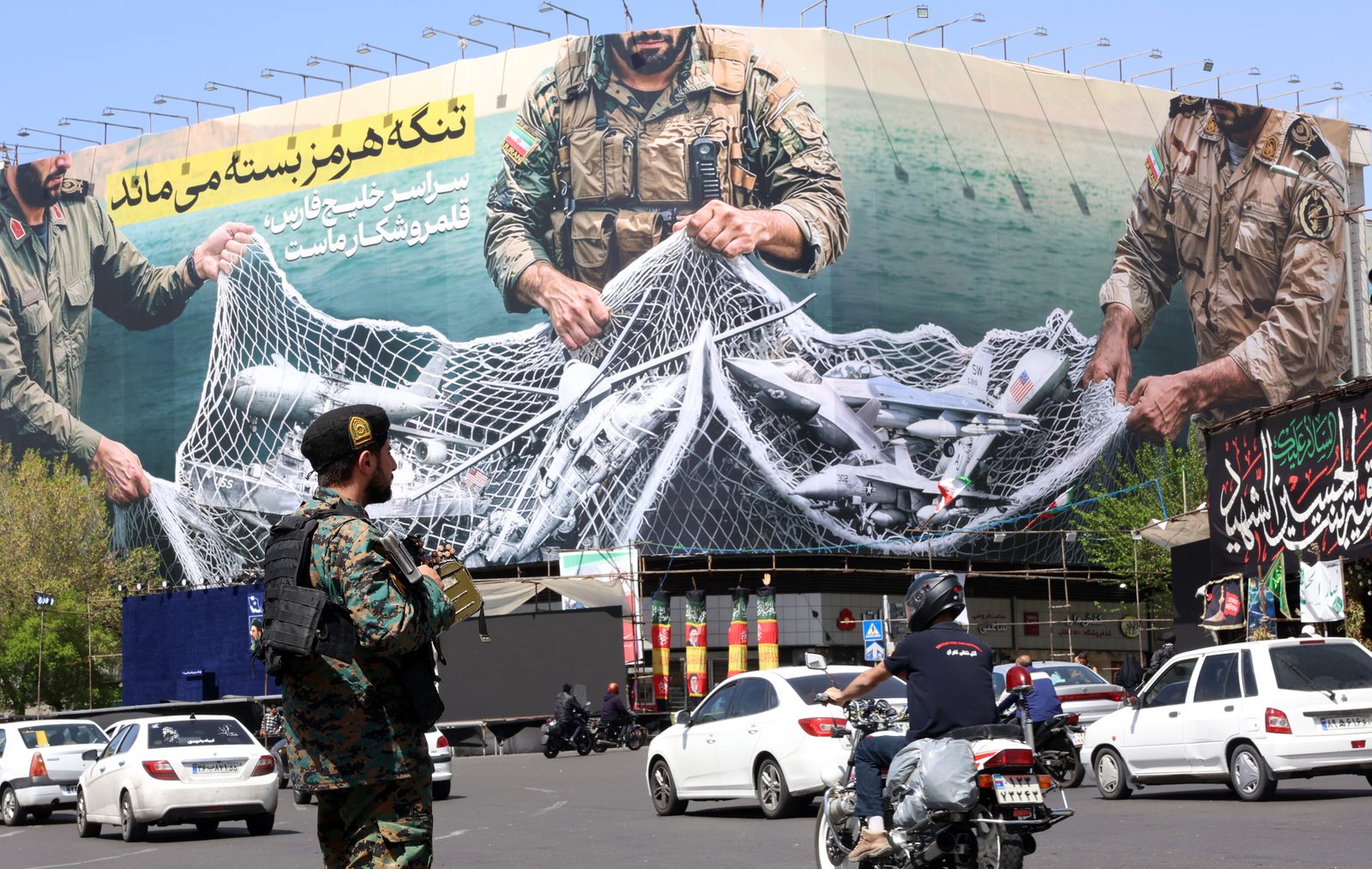 iranian security forces monitor traffic near political billboard stating 'The strait of Hormuz will remain closed'