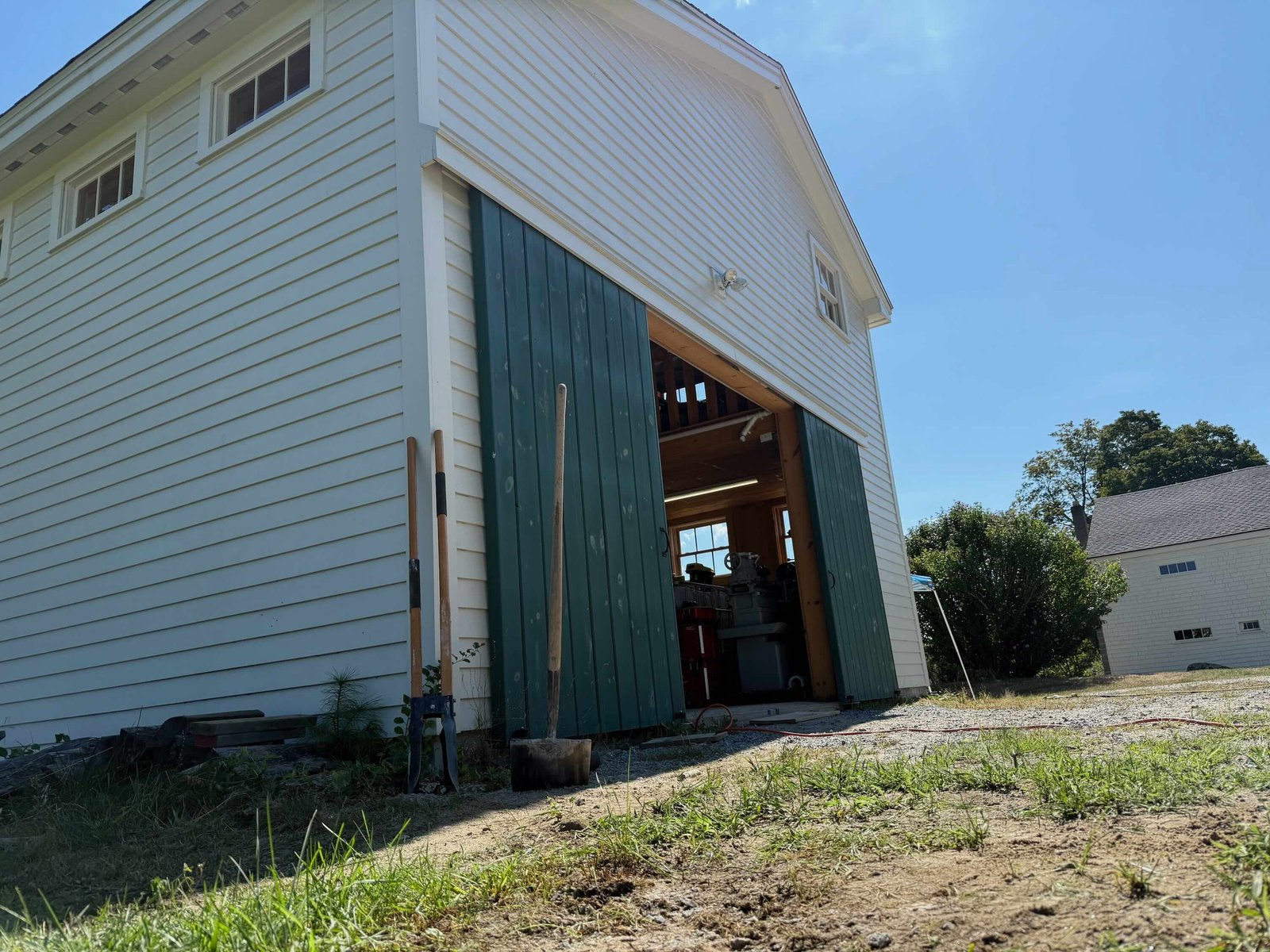 A white shed with green doors casts a shadow on soil and grass.
